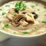 "Bowl of creamy mushroom soup garnished with fresh herbs and served with a slice of crusty bread."