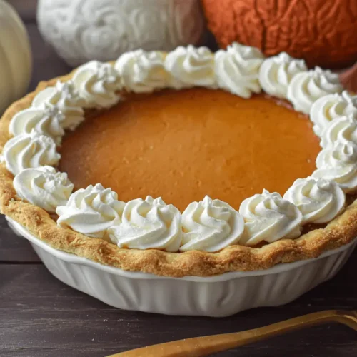 A golden-brown pumpkin pie with whipped cream rosettes and fall decorations on a dark wood surface, showcasing the best pumpkin pie recipe.